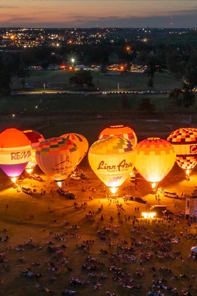 Hot air balloons inflated on a field at dusk with a crowd of people nearby and city lights in the background