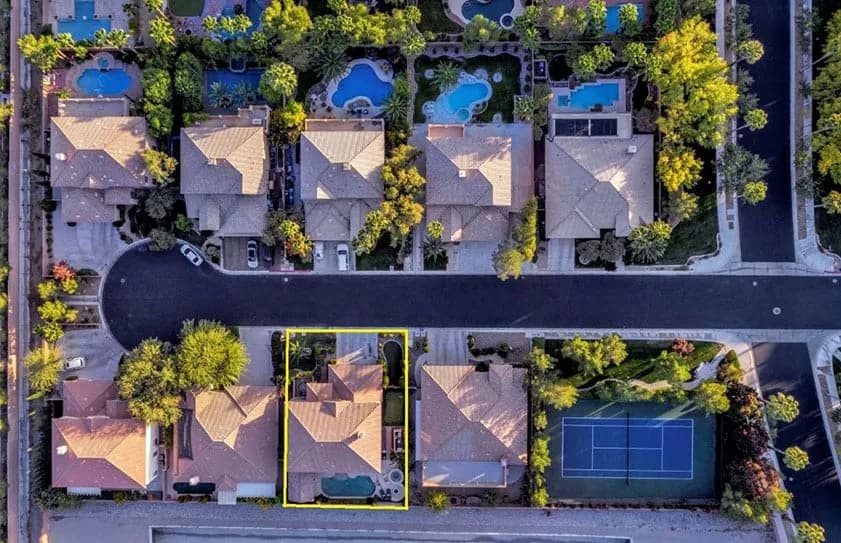 Aerial view of a suburban neighborhood with a yellow-outlined house, pools, and a tennis court