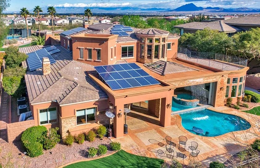 Aerial view of a large stucco house with solar panels, a pool, and a patio with chairs