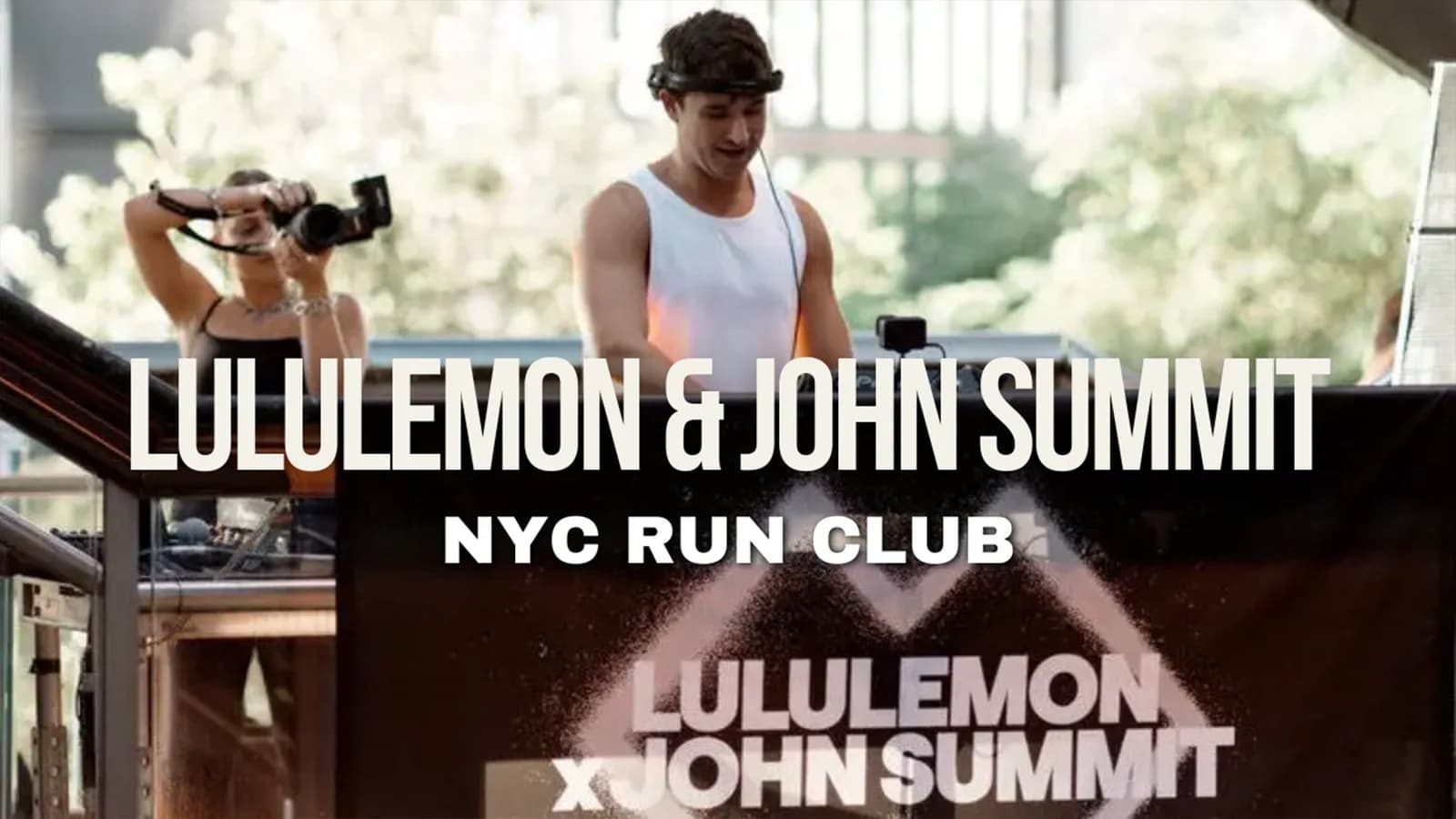 Man in a white tank behind a banner showing Lululemon & John Summit NYC Run Club, a woman photographs him
