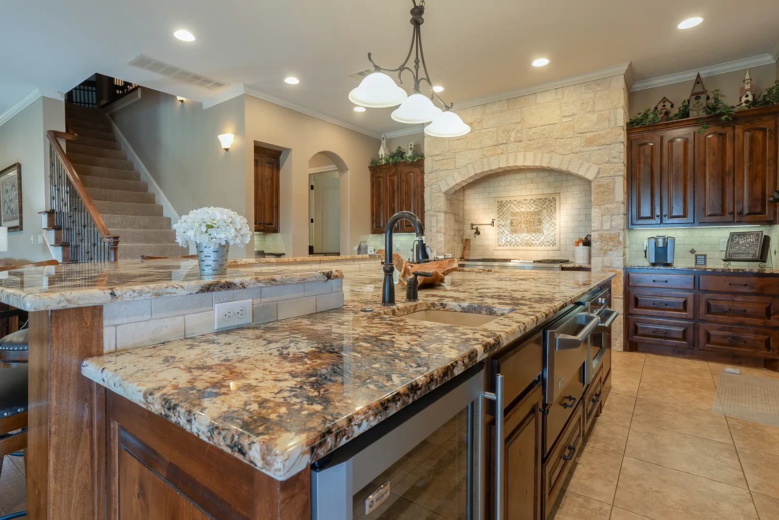 Spacious kitchen with granite island, black faucet, dark wood cabinets, staircase, and stone arched fireplace