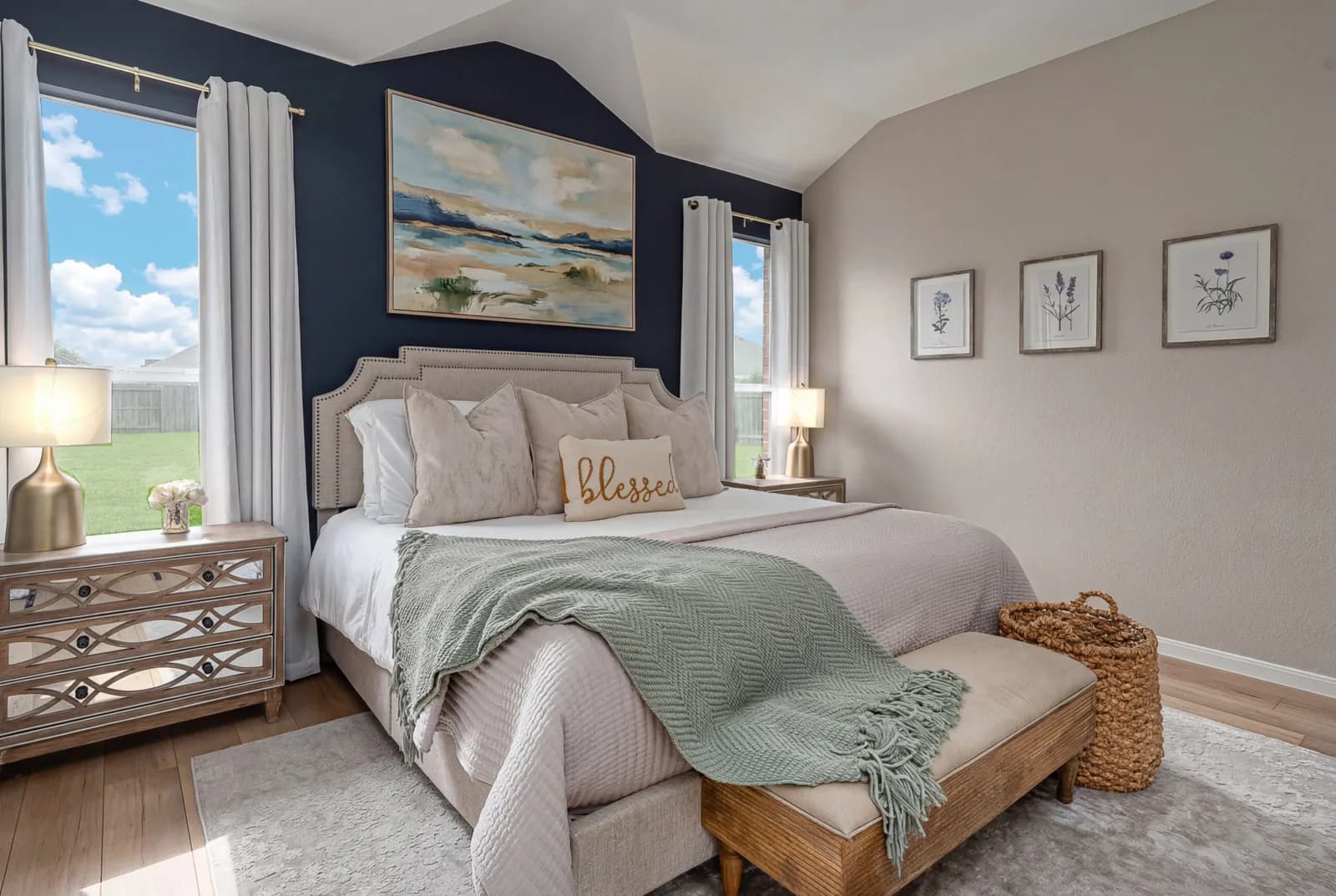 Bedroom with beige upholstered bed against navy wall, framed landscape painting, and two gold bedside lamps