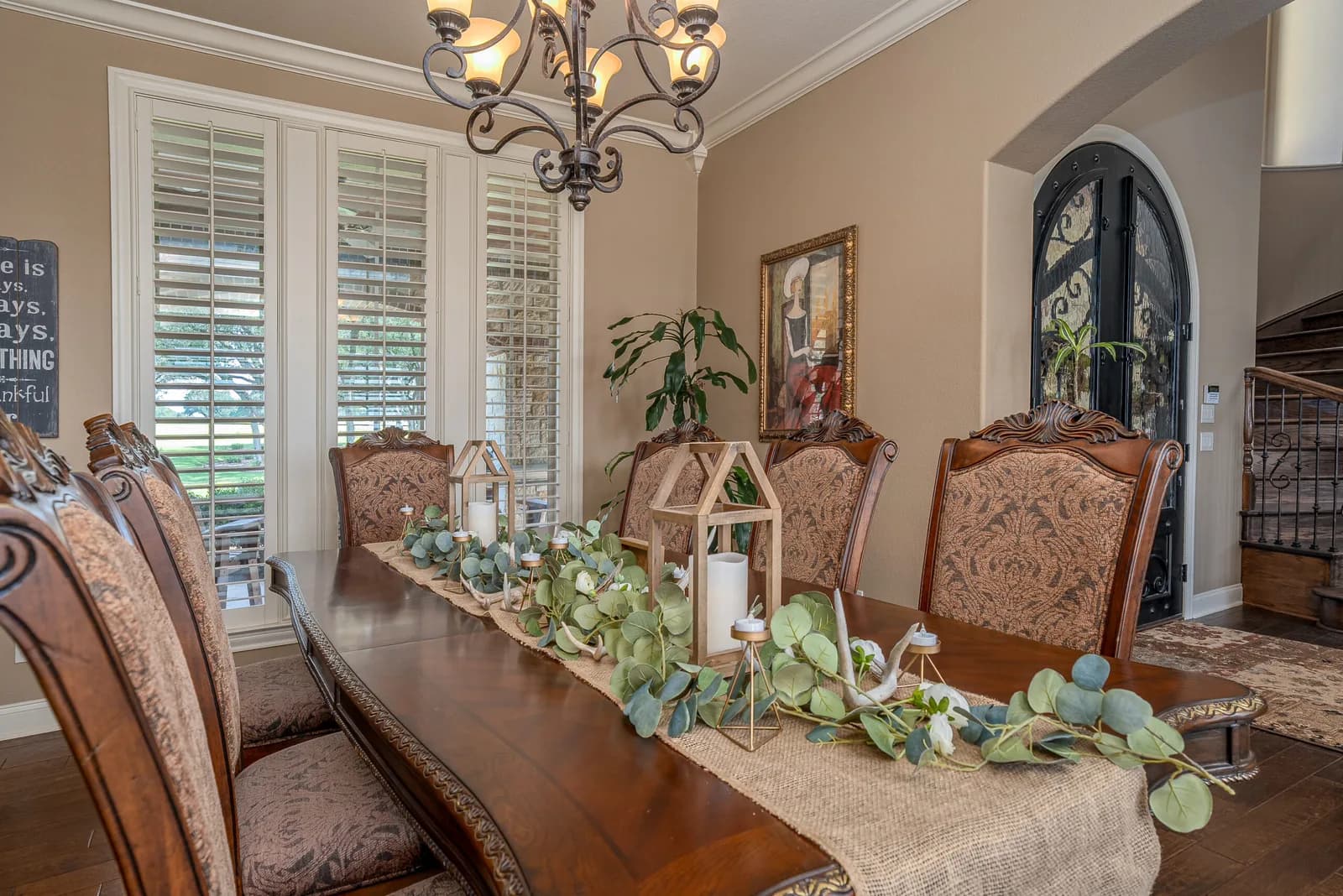 Dining room with a long wooden table, ornate upholstered chairs, eucalyptus centerpiece, chandelier