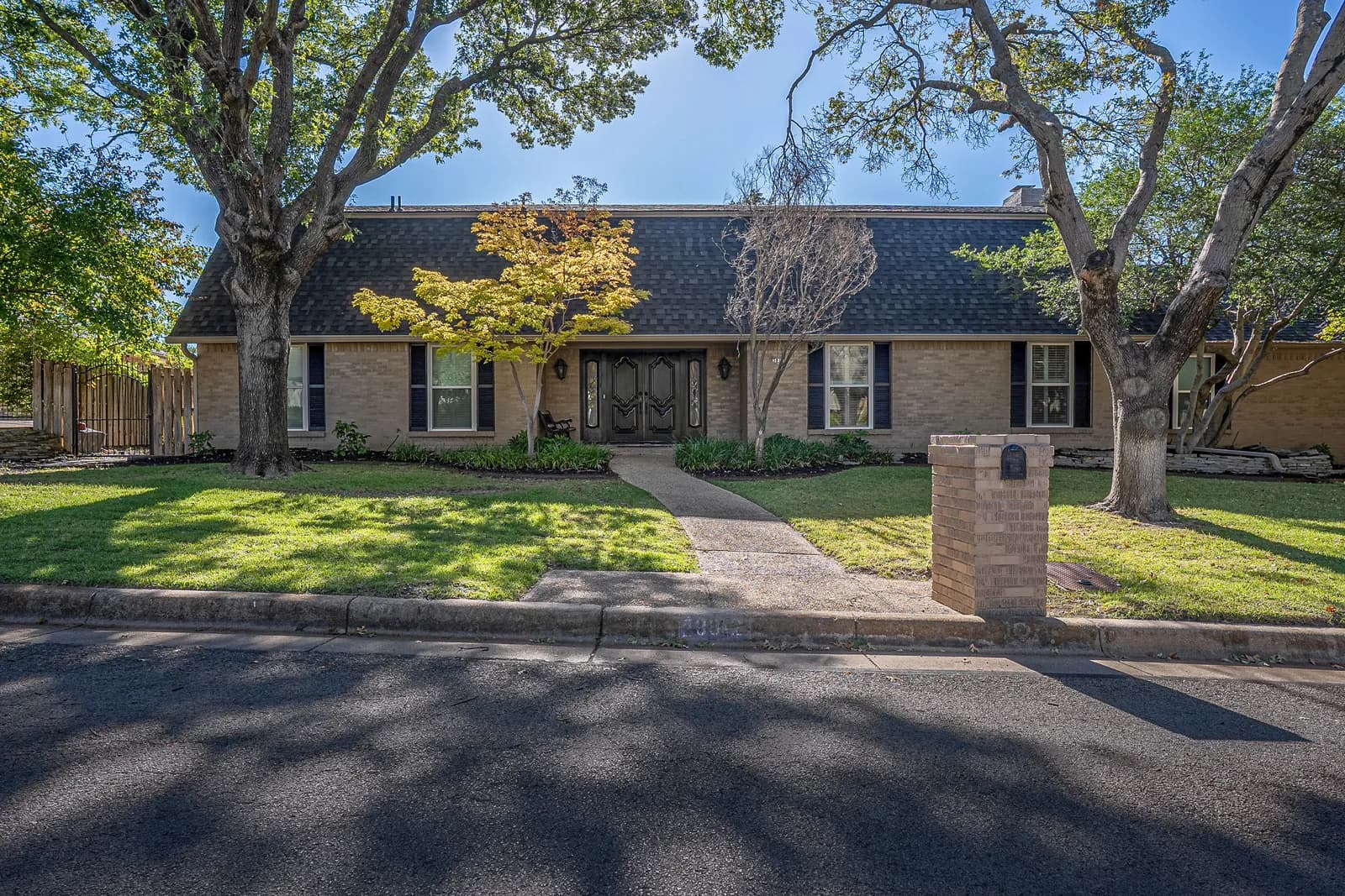 Single-story brick house with dark shingle roof, curved path to double doors, yard trees and brick mailbox