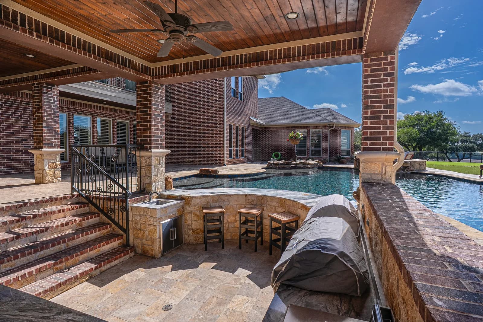 Brick home, covered patio with a ceiling fan, pool with stone bar and stools, and grill