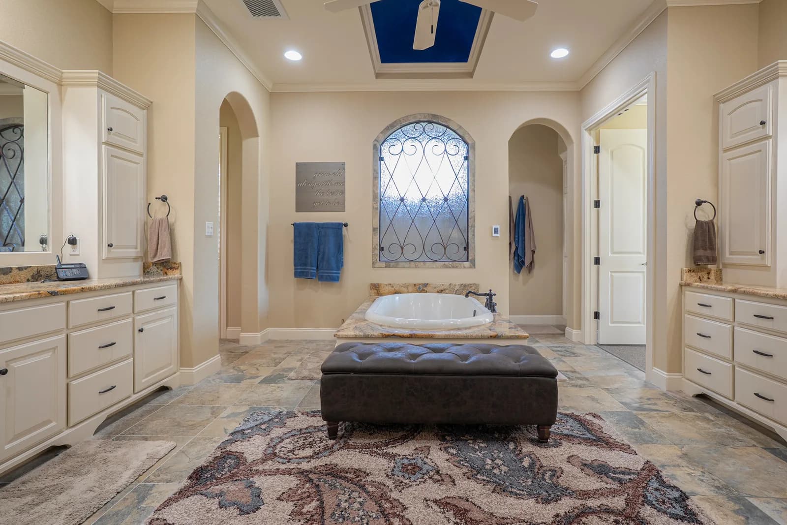 Beige bathroom with oval tub on granite surround, tufted bench, arched window with iron grille, cabinets