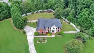 Aerial view of a two-story brick and white house with a curved driveway, green lawn, and surrounding trees