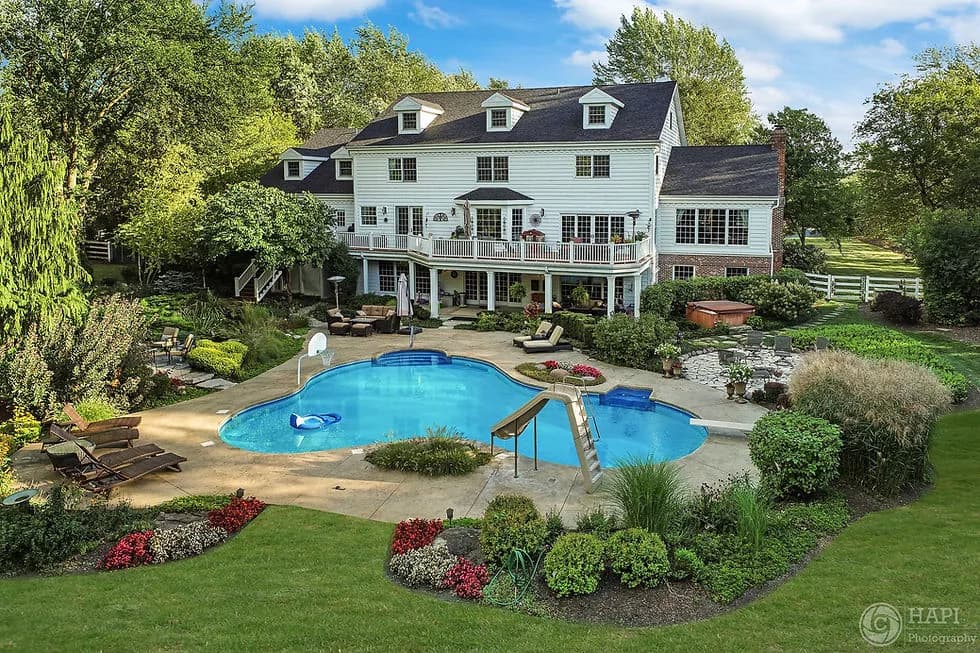 A large white house with a curved pool, deck seating, and lush landscaping in the backyard