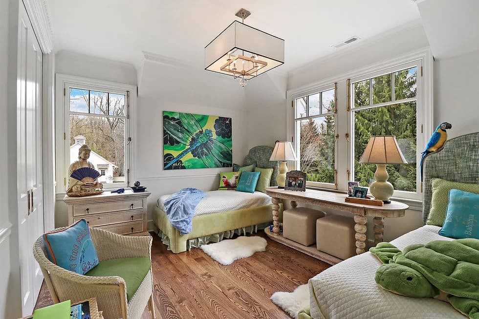 Bright bedroom with two beds, a window-side console table, lamps, artwork, and a green parrot statue