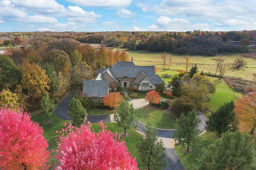 Aerial view of a large house with a circular driveway, surrounded by colorful fall trees and pink foliage