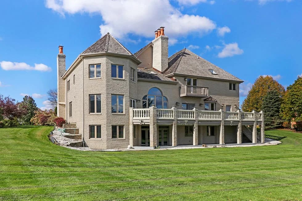 Two-story brick mansion with towers, arched window, and a wraparound balcony on a green lawn