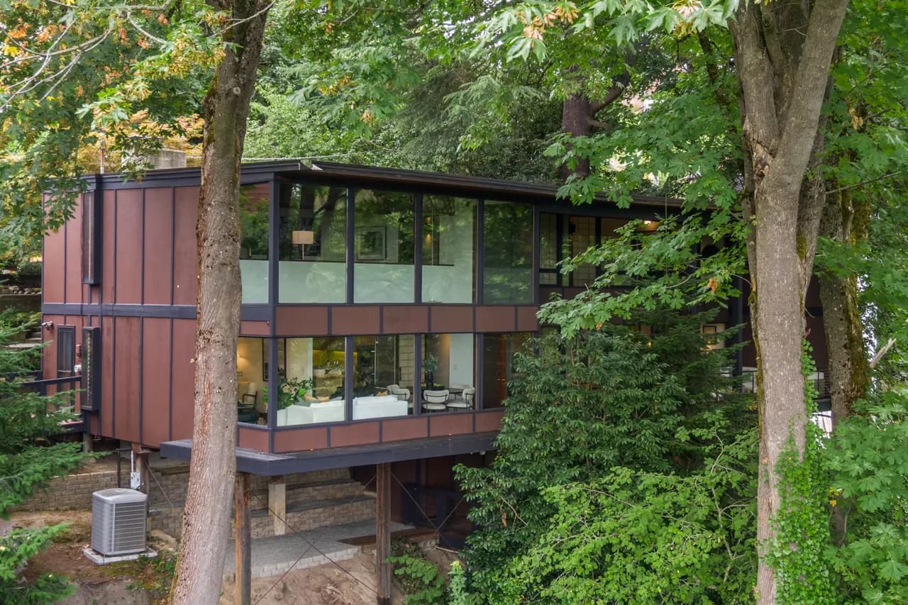 Modern glass-walled two-story house with brown panels, raised on stilts among trees