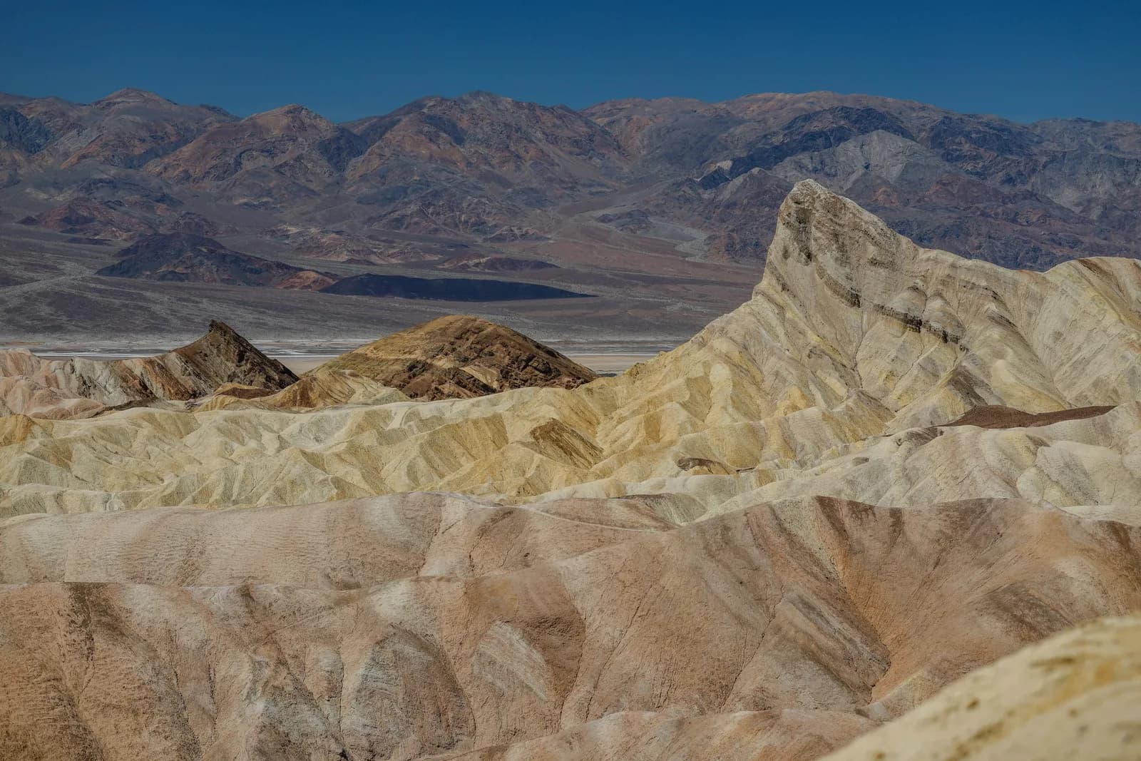 Desert badlands with layered beige and yellow hills, and distant dark mountains under a blue sky