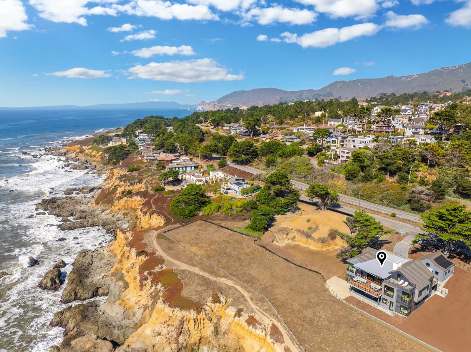 Aerial view of coastal hillside homes with a location pin on a roof, trees, road, rocky shore, and mountains