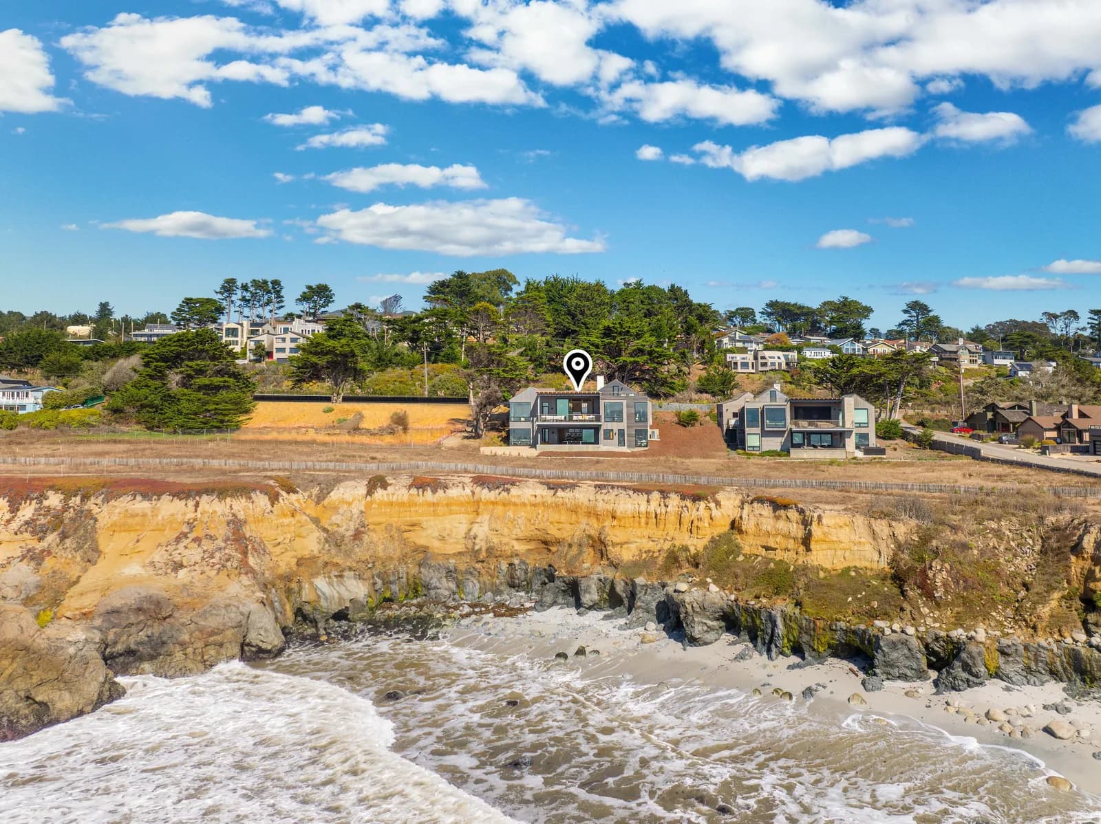 Coastal hillside neighborhood with modern homes on a cliff, waves below, and a location pin over a house