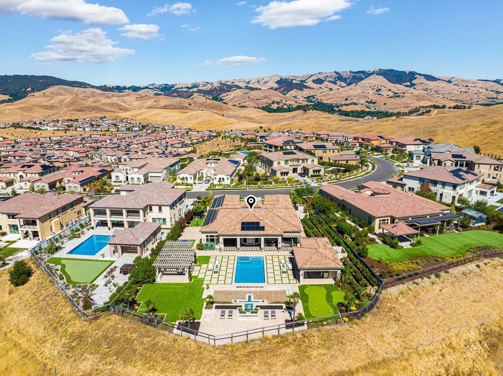Aerial view of beige-roofed houses in a suburb, with pools and manicured lawn, against dry hills
