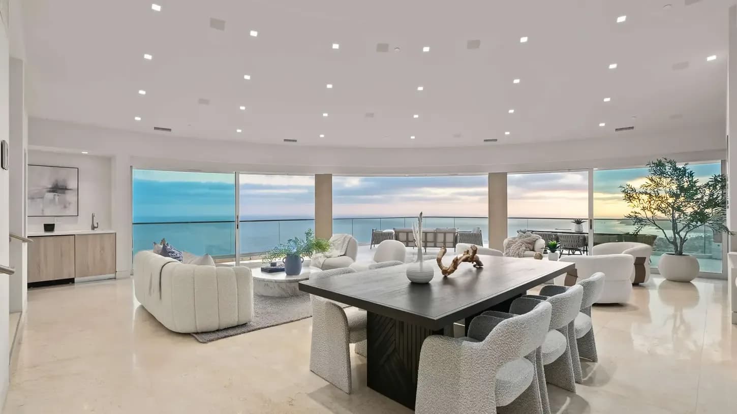 Spacious modern living and dining area with a long wooden table, grey chairs, white sofas, and ocean view
