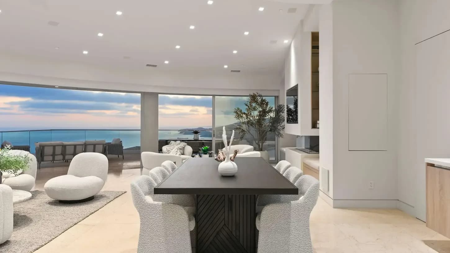 Open living and dining area with a long dark table and light upholstered chairs, and an ocean view