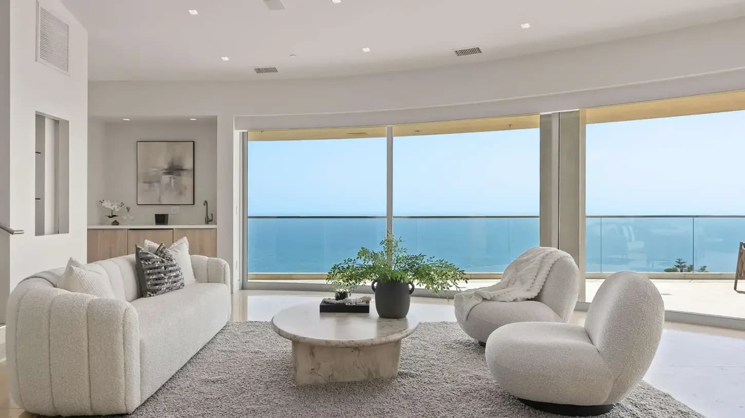 Modern living room with white boucle sofa, curved lounge chairs, marble coffee table, and ocean-view balcony