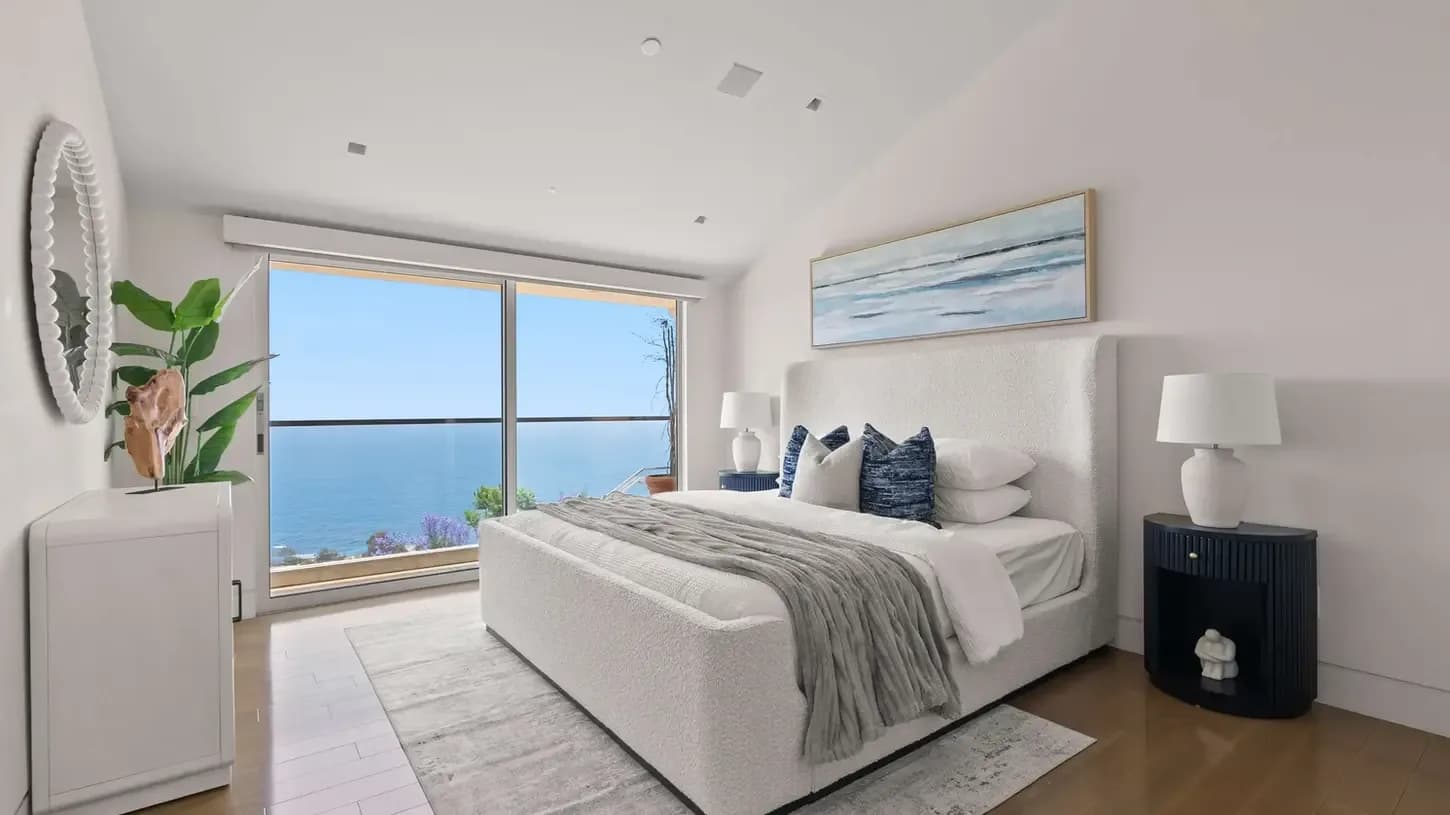 Luxurious white bedroom with a large sea-view window, upholstered bed, blue pillows, and two nightstands with lamps