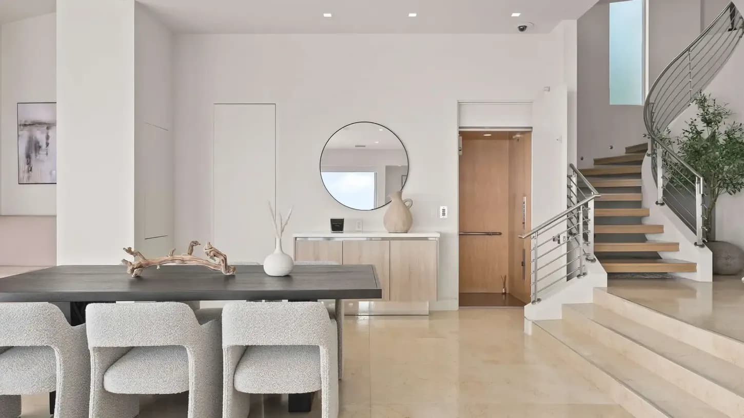 Bright modern interior with a long dark table, driftwood centerpiece, round mirror, and curved staircase