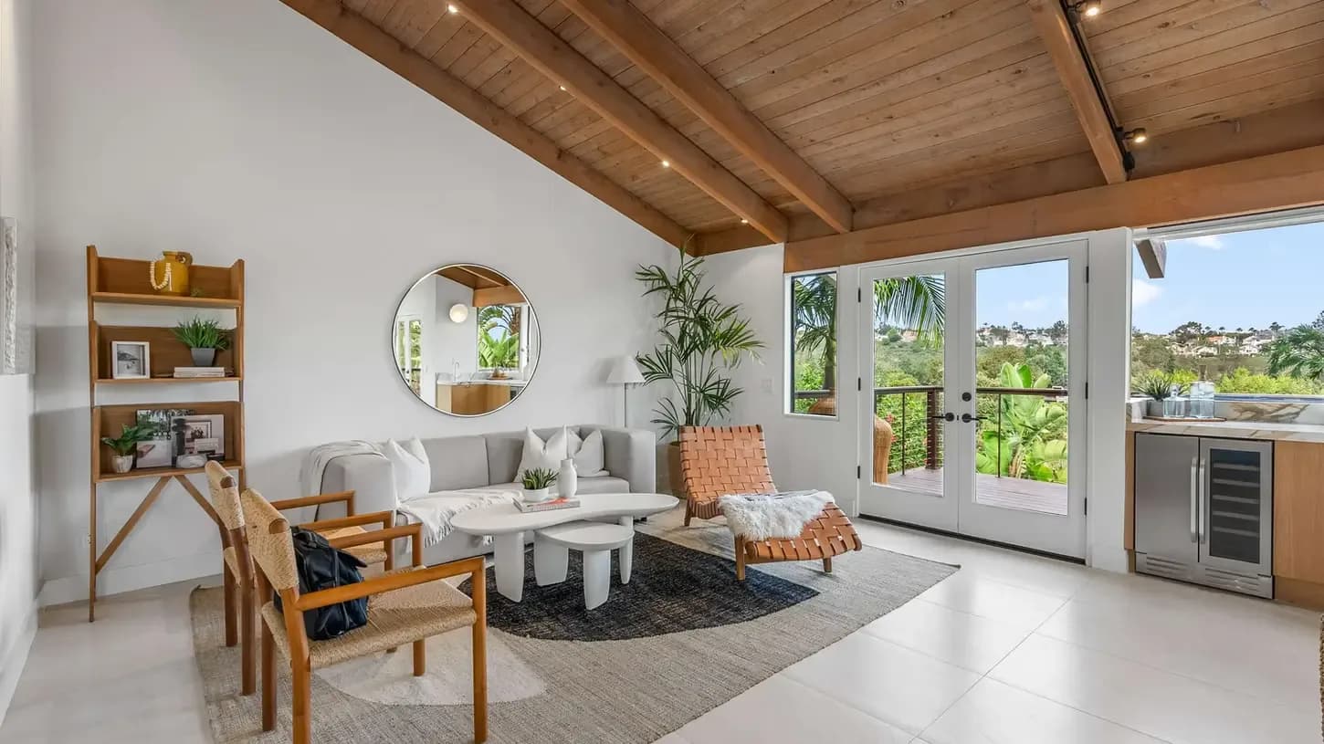 Bright living room with white sofa, wicker chair, round mirror, wooden ceiling, and glass doors to balcony