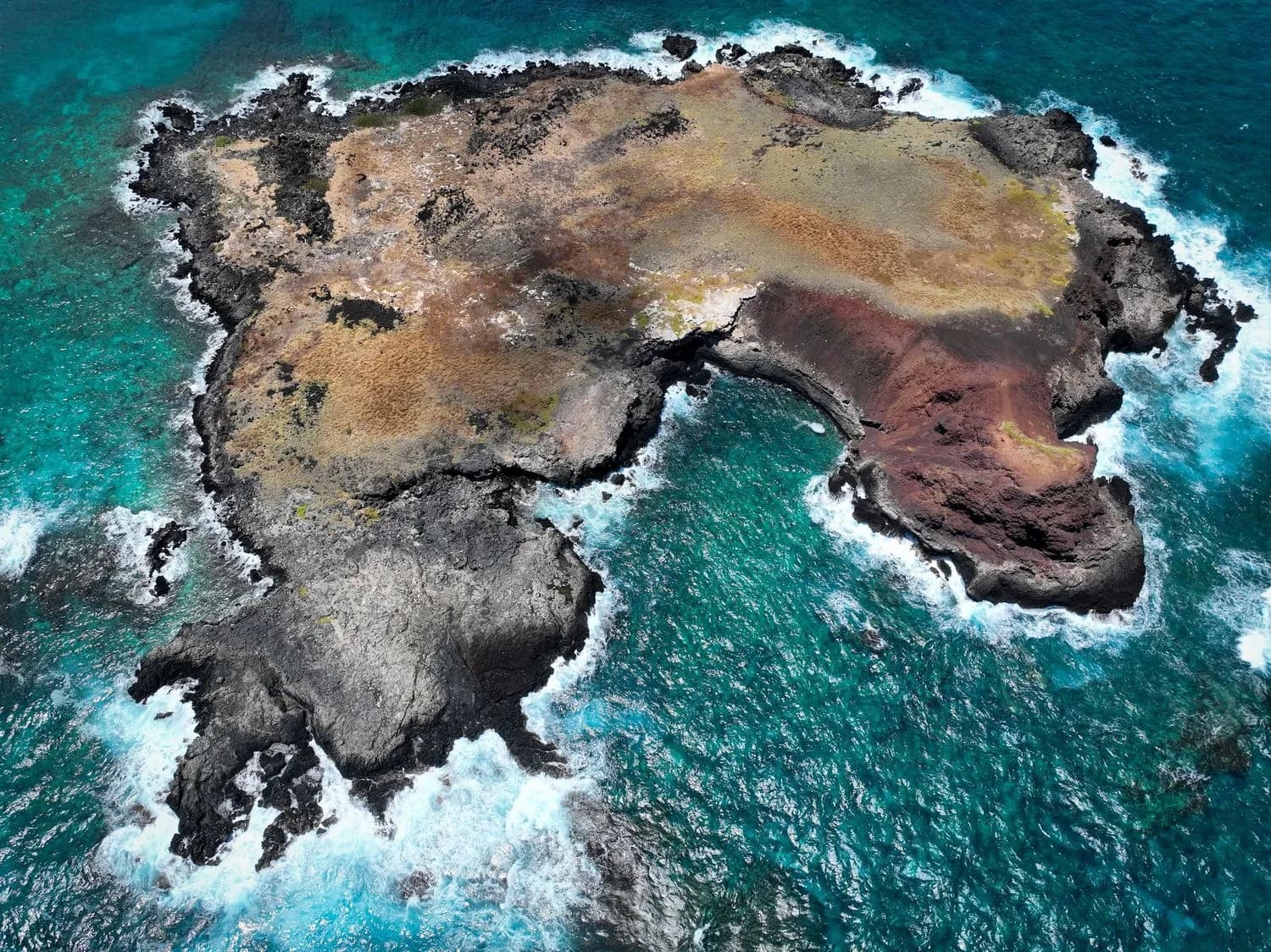 Rocky coastal islet with reddish and black cliffs surrounded by turquoise surf