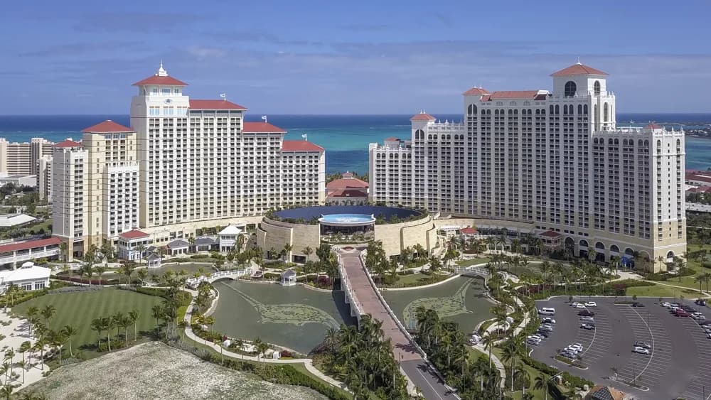 Drone aerial view of a large resort with white towers, red roofs, a central pool, palm trees, and the ocean