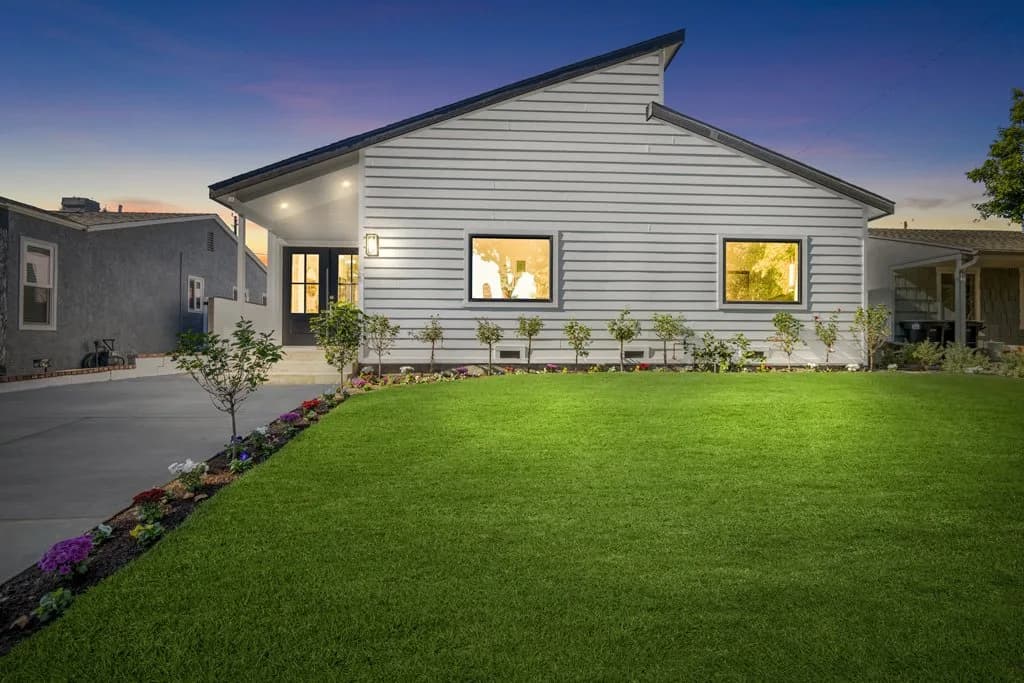 Dusk view of a modern single-story gray house with a green lawn, a flower bed border, and lit windows