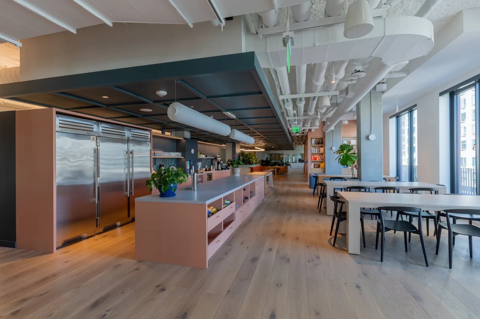 Open-plan space with a long central counter, stainless refrigerators, shelves, seating, and large windows