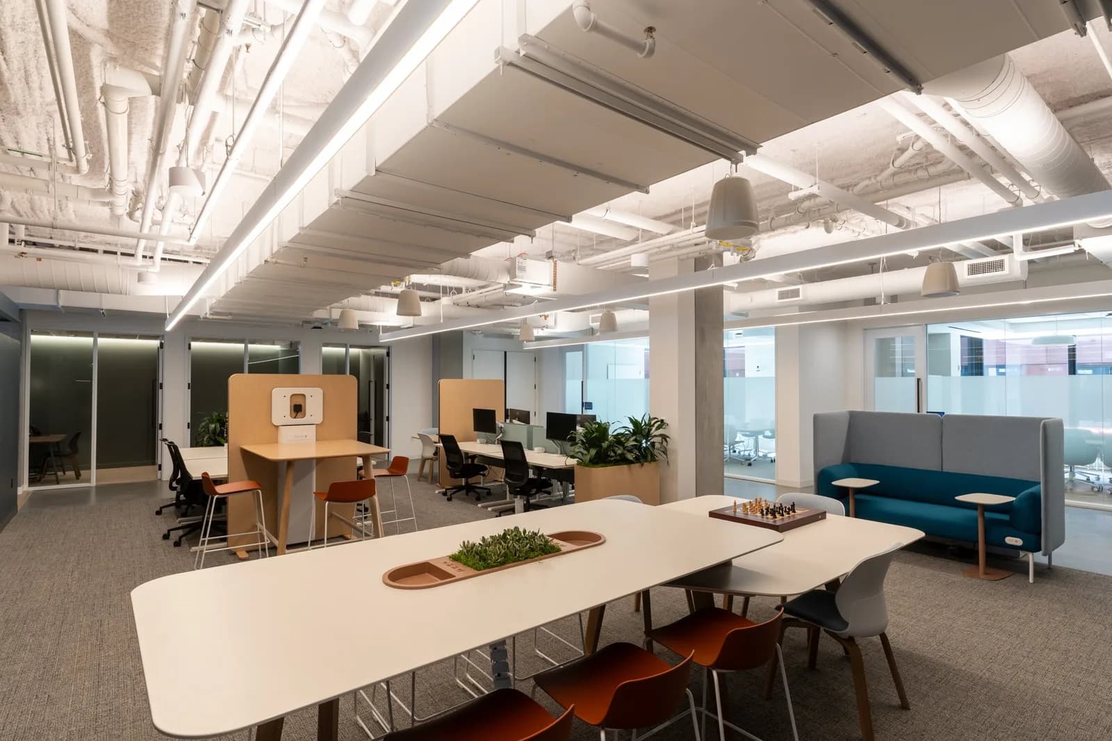 Open-plan office with white tables and gray chairs, chessboard on table, plants and blue-gray seating