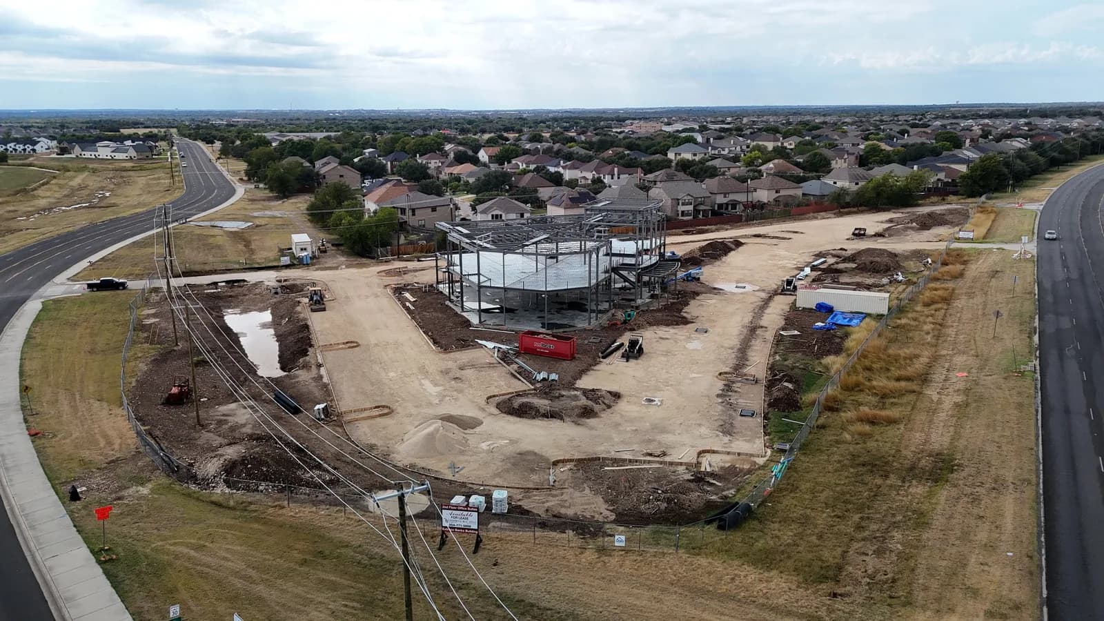 Aerial view of a circular steel-framed building under construction in a fenced site near a residential area