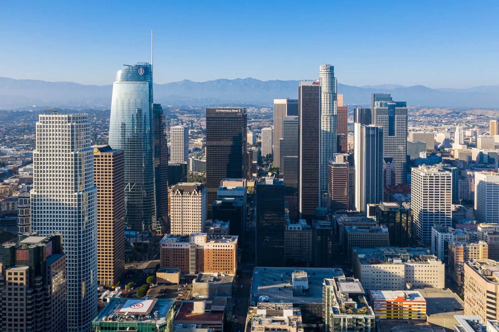 Aerial view of a downtown skyline with glass skyscrapers and distant mountains under a blue sky