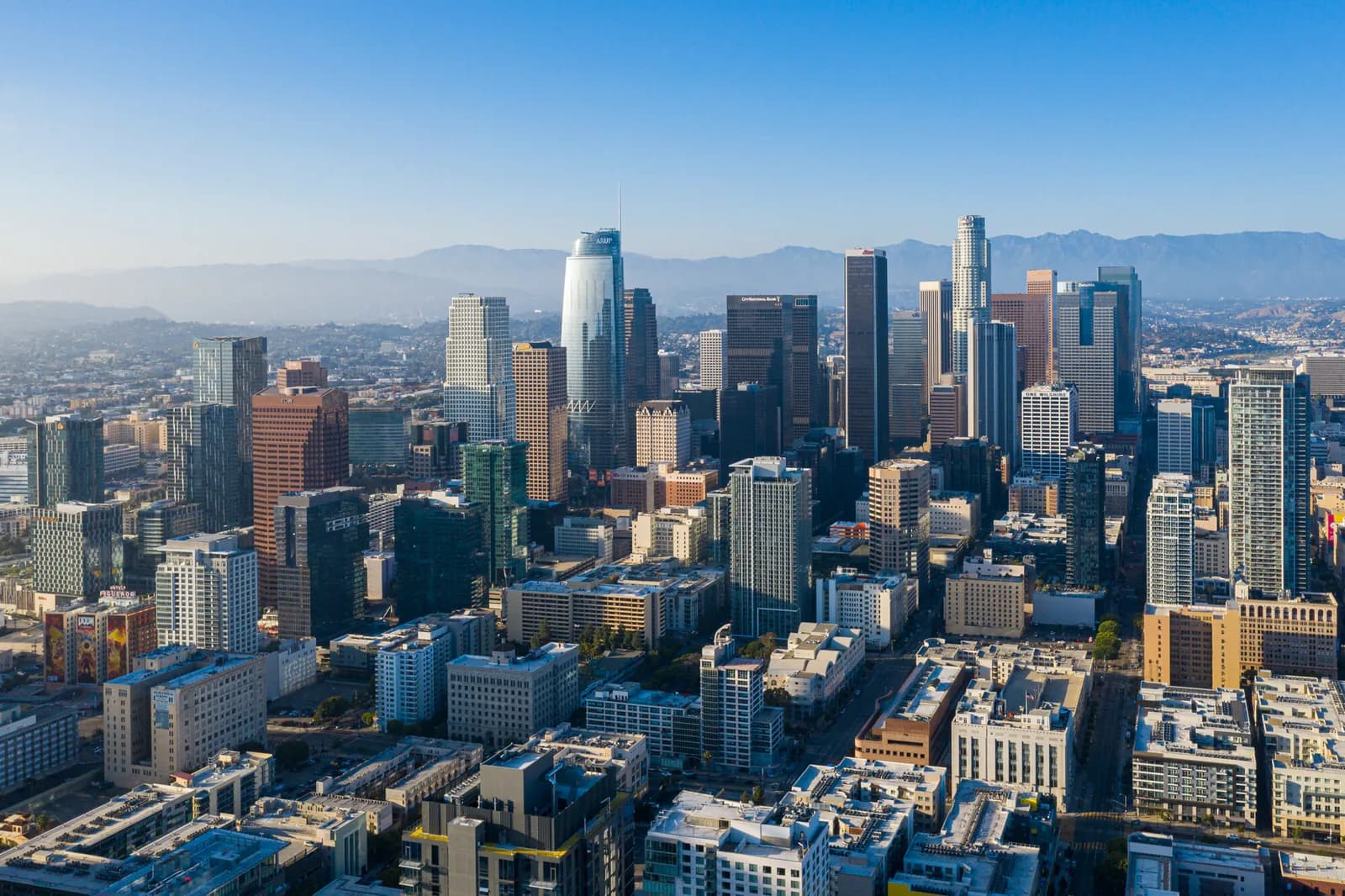 Aerial view of a dense downtown skyline with high-rise buildings and distant mountains under a clear blue sky