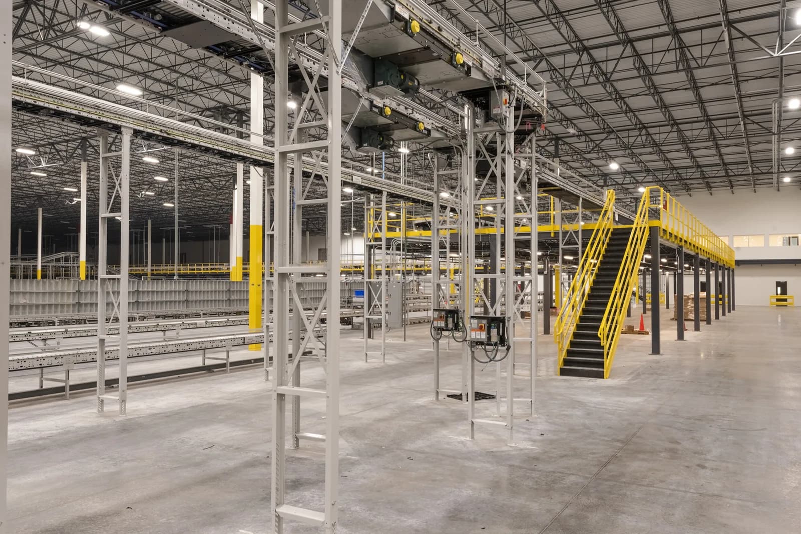 Industrial warehouse interior with gray metal framework, overhead conveyors, and a yellow stairway mezzanine