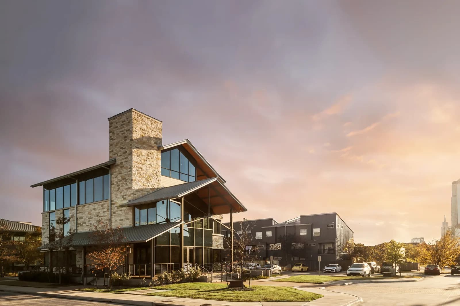 Modern stone building with large glass windows and a tall chimney, cars parked nearby at sunset