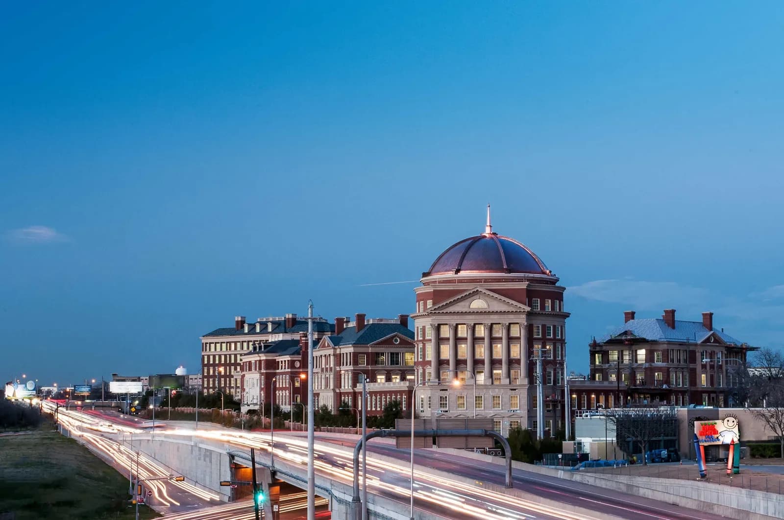 Dusk cityscape with a domed brick building beside a highway, long light trails along the road