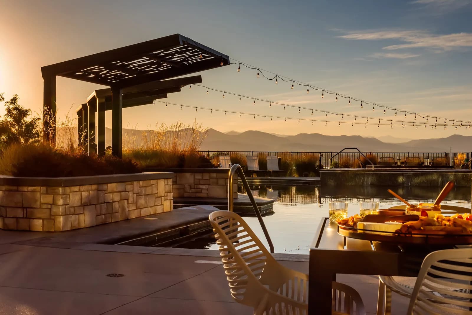 Sunset poolside with string lights, stone planters, lounge chairs, and a food platter on a table