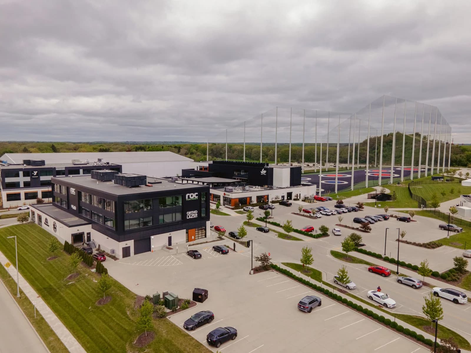 Aerial view of a modern office complex with parking lots and a tall netted driving range on the right
