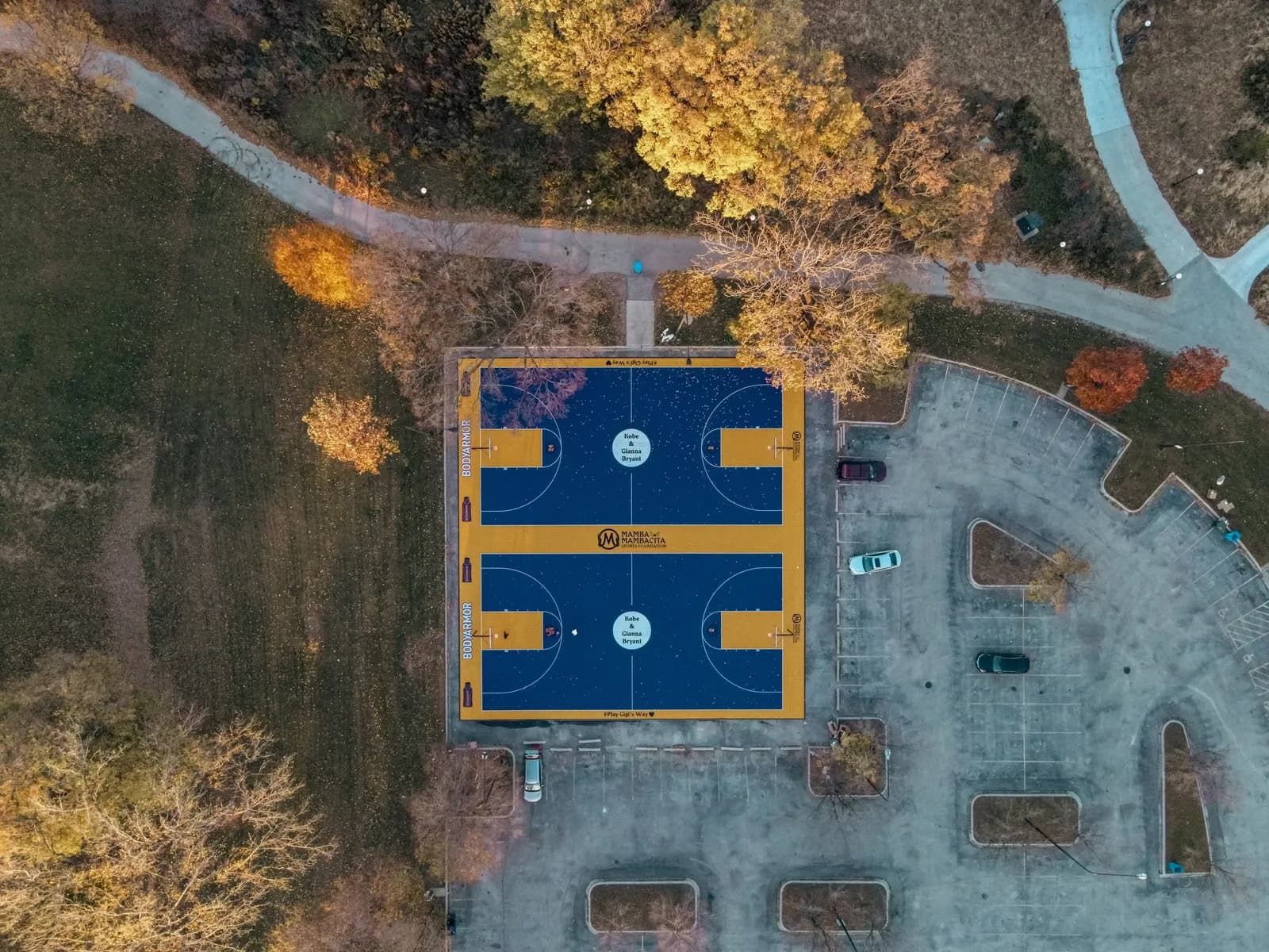 Drone view of two blue basketball courts with yellow borders in a park, autumn trees nearby