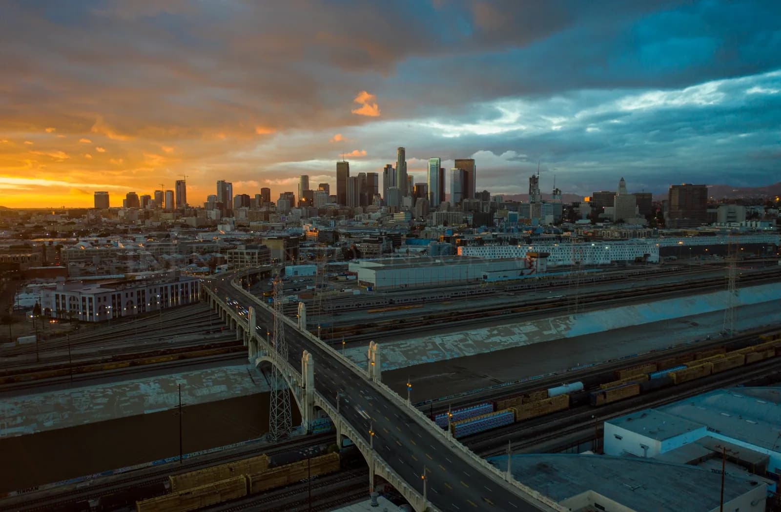 Sunset over a downtown skyline with an arched bridge and rail yard in the foreground