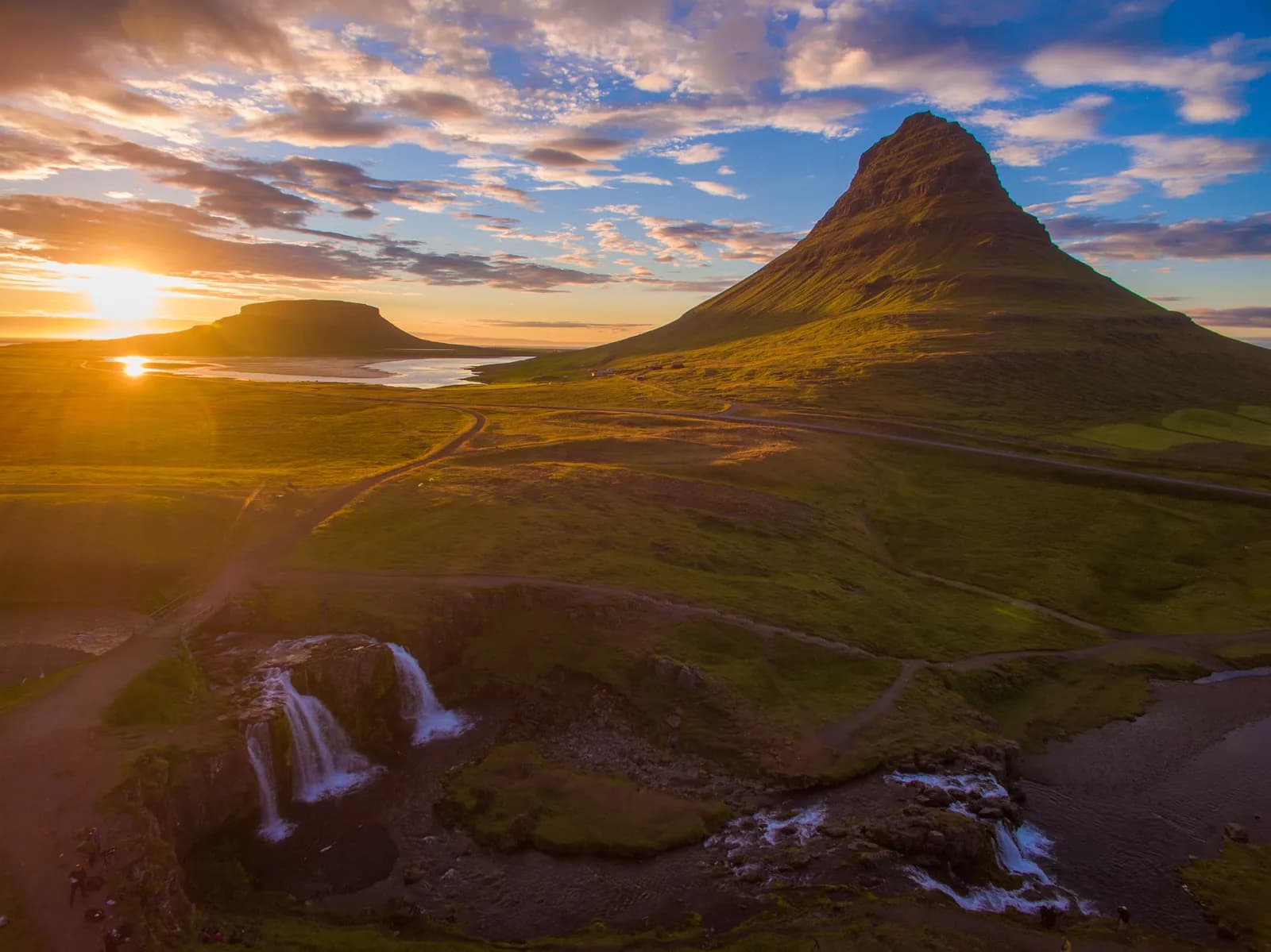Golden hour landscape with a cone-shaped mountain, a small waterfall, and winding dirt paths