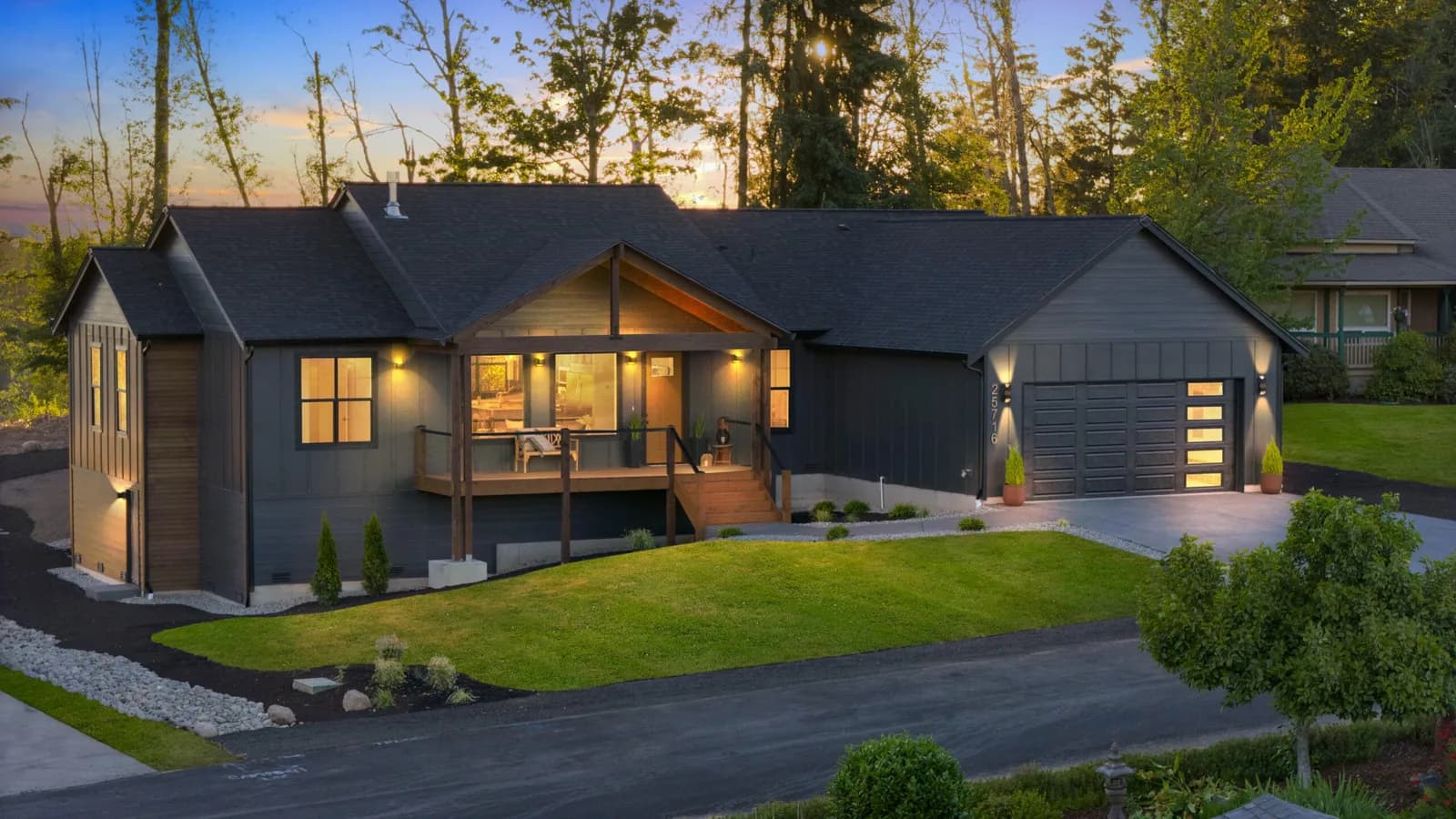 Modern dark gray house with a front porch, two-car garage, warm interior lights, and sunset behind trees