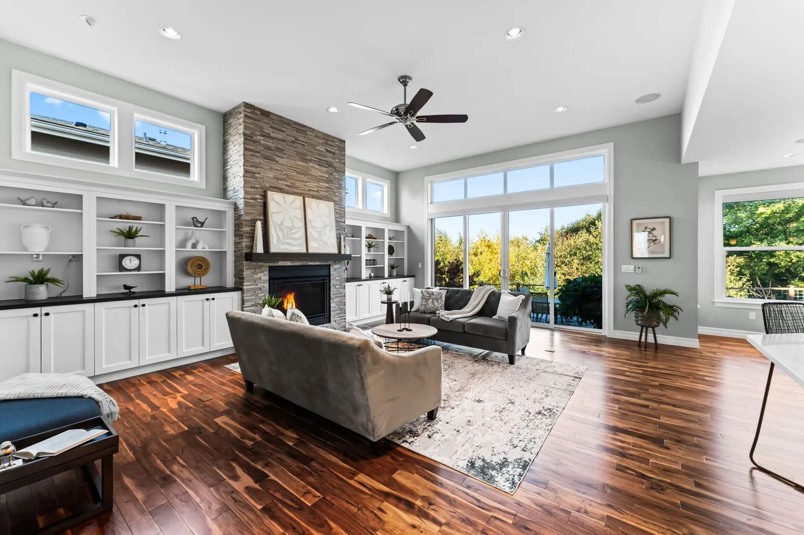 Spacious living room with stone fireplace, white built-in shelves, hardwood floors, and large windows