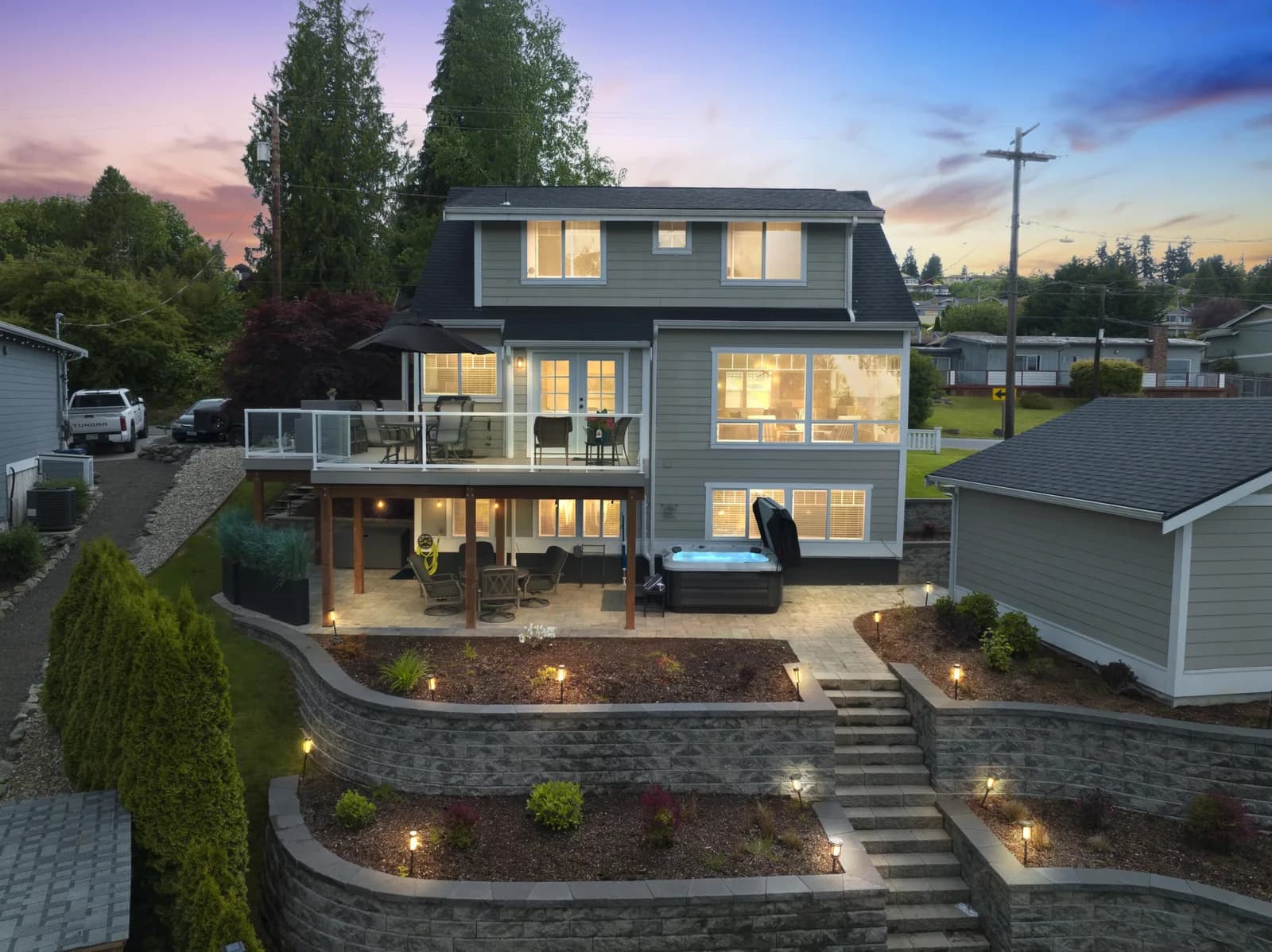 Aerial view of a gray two-story house with a glass-railed deck, patio, hot tub, and terraced garden at sunset