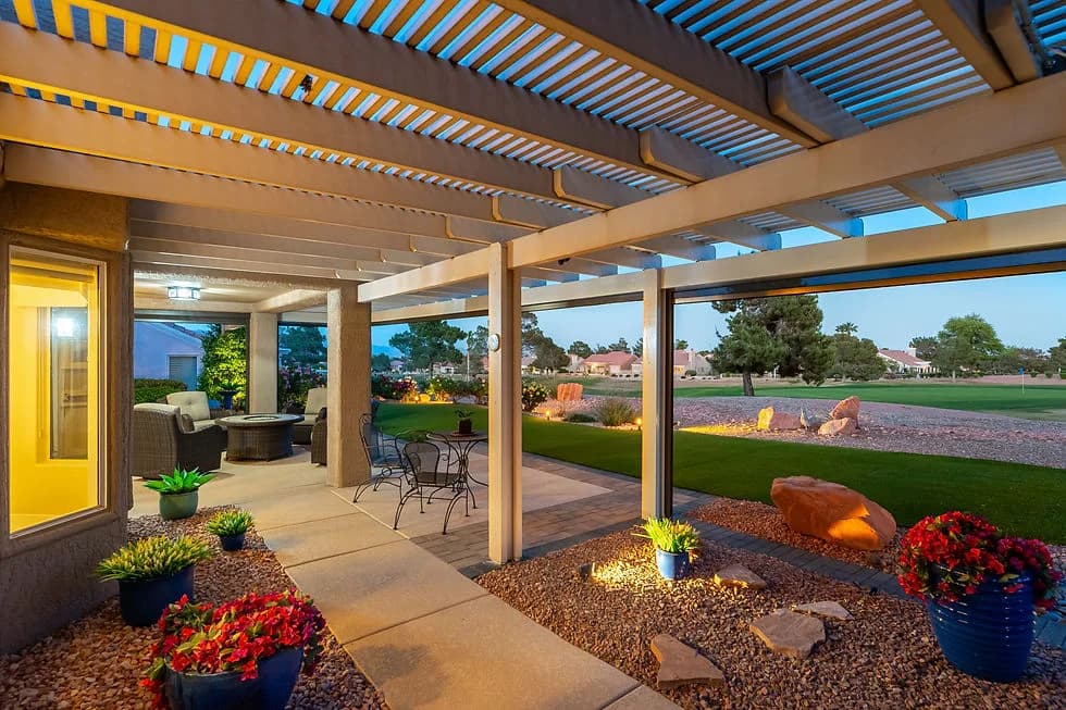 Patio under a slatted pergola with outdoor seating, potted plants, gravel, and a lawn beyond