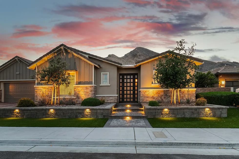 Front entrance to a modern single-story home with stone accents, lighting, trees, and mountains at sunset