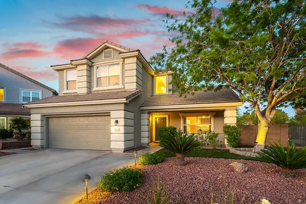 Two-story beige house with a two-car garage, front porch lights, and a tree on the right