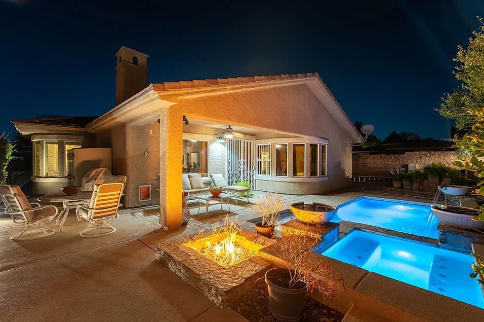 Nighttime backyard with a lit stone fire pit, blue illuminated pool, and patio seating beside a stucco house