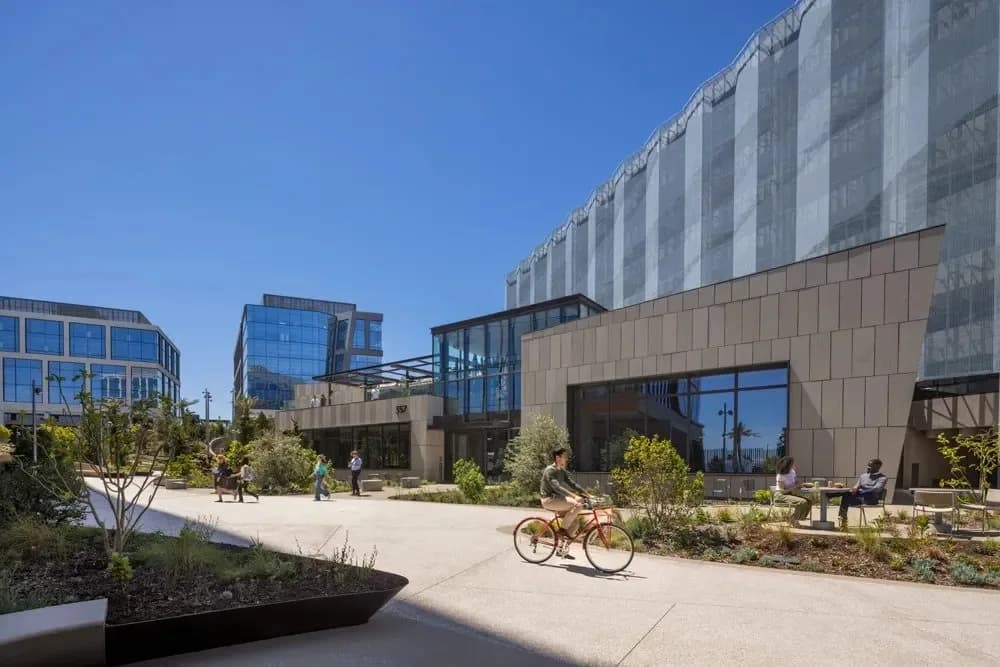 A cyclist on a red bike rides along a concrete path past glass office buildings in a modern campus courtyard